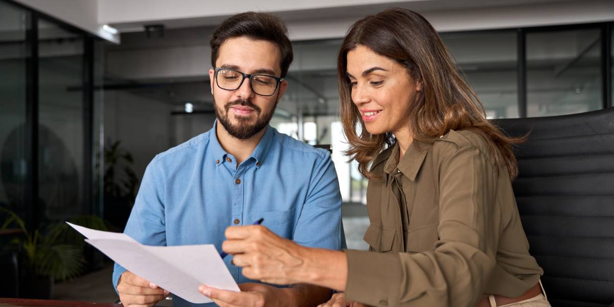 Dois profissionais em um ambiente de escritório moderno, analisando juntos um documento. O homem, com barba, óculos e camisa azul, segura o papel enquanto a mulher ao lado, vestindo uma blusa verde-oliva, aponta algo no documento com um sorriso. A cena representa uma colaboração ou revisão de trabalho, transmitindo uma atmosfera de trabalho em equipe e cooperação, currículo