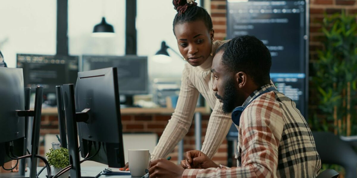 Dois programadores colaborando em um ambiente de escritório moderno. A mulher está em pé ao lado do colega, discutindo um projeto exibido no monitor do computador. O ambiente apresenta uma decoração industrial com detalhes em tijolo aparente e plantas, refletindo um espaço de trabalho colaborativo e tecnológico (trabalho, cultura, time, pessoas, OutSystems, programadores, desenvolvedores, oportunidades, tecnologia, salarios, salario)