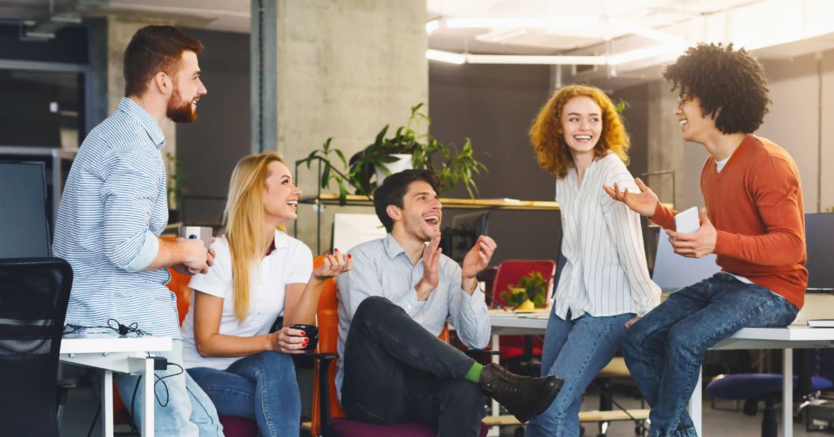 Cinco pessoas estão reunidas em um ambiente de escritório moderno, descontraído e iluminado. Três homens e duas mulheres sorriem e conversam animadamente. Eles estão sentados ou encostados em cadeiras, vestindo roupas casuais, e há plantas e mesas ao fundo, contribuindo para uma atmosfera acolhedora e colaborativa.