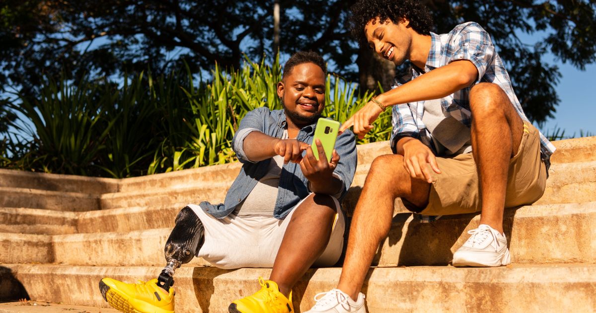 A imagem mostra dois amigos sentados em degraus ao ar livre num dia ensolarado, rodeados por vegetação verde. Ambos estão sorrindo enquanto olham para um telemóvel na mão do amigo à esquerda, que tem uma prótese na perna e está a usar ténis amarelos brilhantes e roupa casual. O outro amigo, também vestido de forma descontraída, aponta para o ecrã do telemóvel com entusiasmo. A cena transmite uma sensação de amizade, inclusão