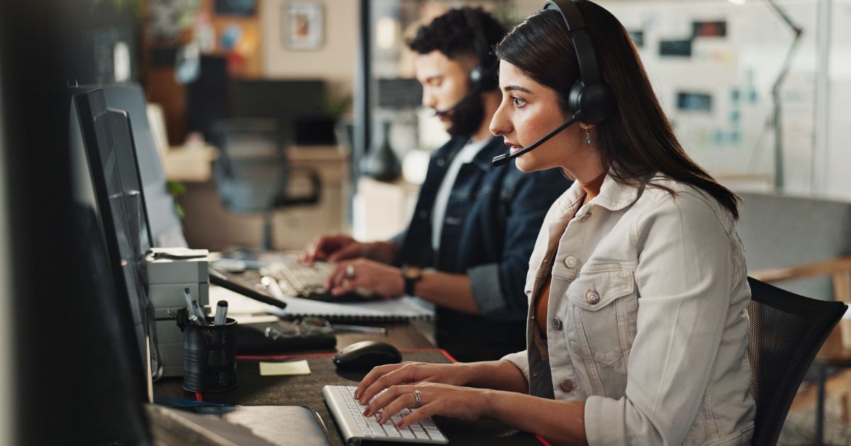 Uma mulher jovem com cabelos castanhos longos e pele clara está sentada em uma estação de trabalho, usando um headset preto com microfone, digitando em um teclado branco. Ela está focada na tela de um computador, com uma expressão séria e concentrada. Ela veste uma jaqueta bege casual. Atrás dela, há um homem também usando headset e trabalhando no computador, ligeiramente desfocado. O ambiente é um escritório moderno com luz natural e uma atmosfera profissional. Na mesa, há canetas em um suporte, papéis e outros objetos de trabalho.