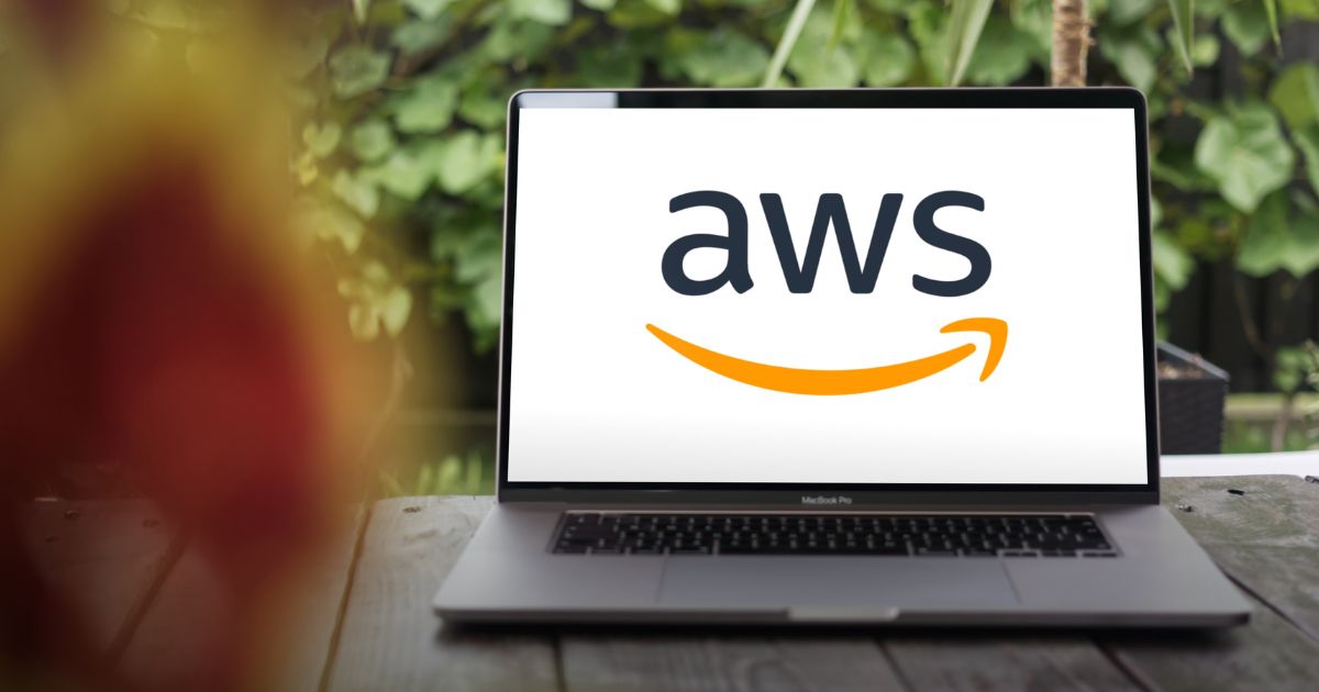 Um computador portátil com a marca MacBook Pro está sobre uma mesa ao ar livre. O ecrã mostra o logótipo da AWS (Amazon Web Services), com letras pretas e o ícone de uma seta curva laranja por baixo, que também remete para um sorriso. Ao fundo, há um ambiente natural com folhas verdes desfocadas, sugerindo um cenário tranquilo ou de trabalho remoto.