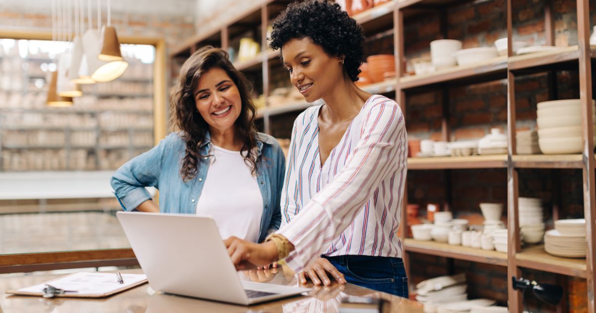 Duas mulheres sorridentes em um ambiente de loja de cerâmica. Elas estão em pé ao lado de uma bancada, olhando para um laptop. Ao fundo, prateleiras exibem tigelas, pratos e outros itens de cerâmica. A cena sugere colaboração e trabalho em equipe em um ambiente criativo e acolhedor.