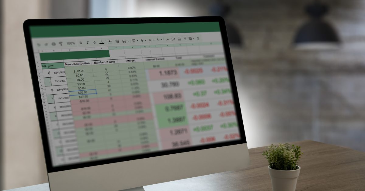 Monitor de computador exibindo uma planilha com dados financeiros em destaque, incluindo colunas de contribuições, número de dias, juros e totais. Uma pequena planta decorativa está posicionada sobre a mesa ao lado do monitor, em um ambiente de trabalho moderno e bem iluminado.