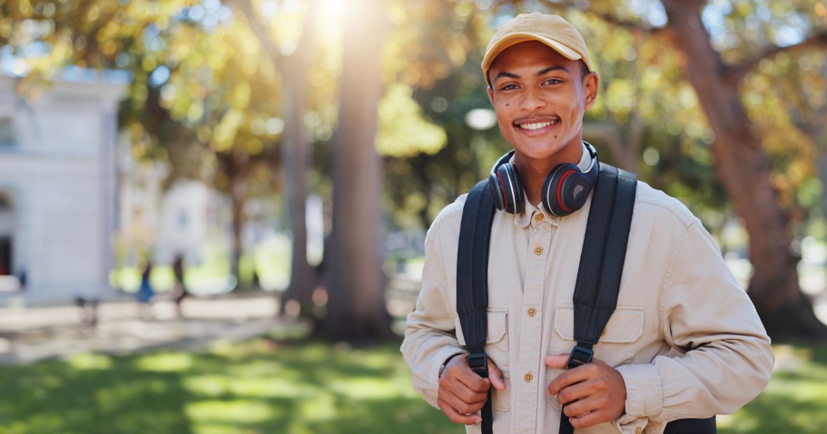 Um jovem sorridente com pele morena e um rosto alegre está ao ar livre em um ambiente arborizado, possivelmente um campus universitário ou parque. Ele usa um boné amarelo, uma camisa bege e carrega uma mochila preta nos ombros. Um par de fones de ouvido pretos e vermelhos descansa ao redor do seu pescoço. O fundo é desfocado, com árvores, gramado verde e algumas pessoas caminhando ao longe sob a luz do sol.