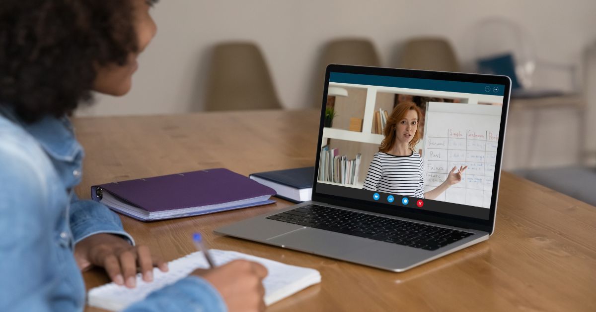 Uma jovem estudante, vestindo uma jaqueta jeans, está sentada à mesa com um caderno aberto e caneta na mão, assistindo a uma aula online em seu laptop. Na tela do computador, uma professora ruiva, vestindo uma blusa listrada, aponta para um quadro branco com anotações. Ao fundo, há uma estante com livros e objetos decorativos. A cena transmite um ambiente de aprendizado remoto e concentração.