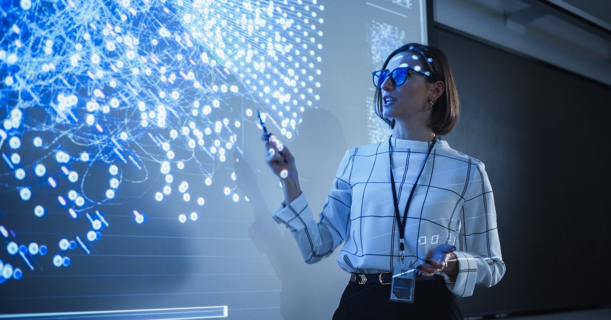 Uma mulher usando óculos escuros e uma blusa branca com padrão quadriculado apresenta um gráfico de dados projetado em uma tela grande. Ela segura uma caneta apontadora e um dispositivo eletrônico, gesticulando enquanto explica as informações. O gráfico projetado exibe uma complexa rede de pontos e conexões azuis brilhantes, sugerindo um tema relacionado a tecnologia, ciência de dados ou inteligência artificial. O ambiente tem iluminação baixa, destacando a projeção e criando um efeito futurista.