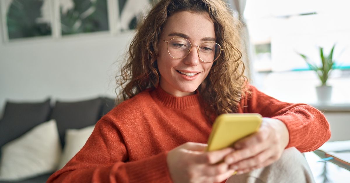 Uma mulher jovem com cabelos cacheados castanhos, pele clara e óculos de armação fina está sentada em um ambiente aconchegante. Ela usa um suéter laranja e segura um smartphone amarelo, sorrindo enquanto olha para a tela. Ao fundo, há um sofá cinza com almofadas claras, algumas plantas e quadros decorativos na parede. A cena é iluminada por luz natural, transmitindo uma sensação de conforto e tranquilidade.